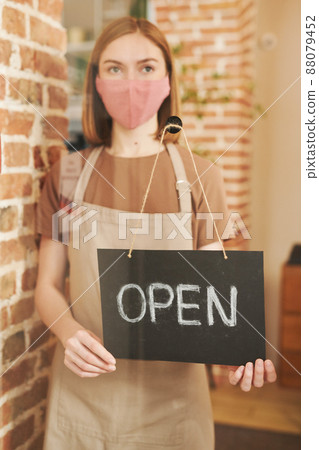 Vertical through glass door portrait of young female waitperson wearing protective mask on face starting workday with opening cafe 88079452