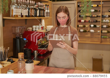 Vertical medium long shot of modern female cafe worker wearing protective mask standing at counter checking order using digital tablet Vertical medium long shot of modern female cafe worker wearing protective mask standing at counter checking order using digital tablet 88079455