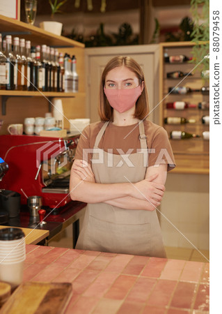 Vertical medium long shot of modern female waitperson wearing protective mask standing at counter with arms crossed looking at camera 88079458