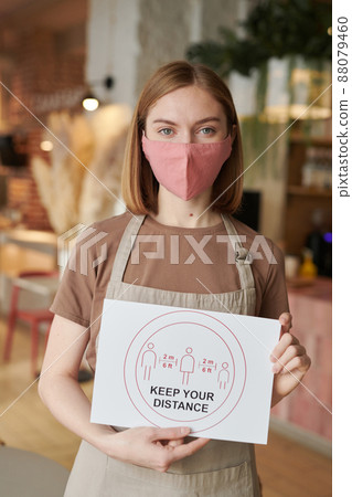 Vertical medium portrait shot of unrecognizable waitperson wearing apron standing in cafe holding Keep Your Distance sign looking at camera Vertical medium portrait shot of unrecognizable waitperson wearing apron standing in cafe holding Keep Your Distance sign looking at camera 88079460