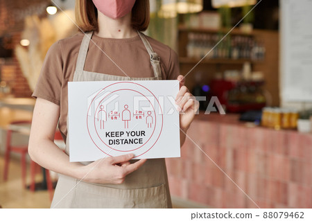 Horizontal medium section shot of unrecognizable waitperson standing in cafe holding Keep Your Distance sign 88079462