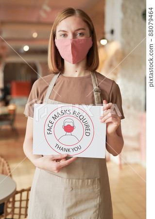 Vertical shot of unrecognizable female cafe worker standing alone holding Face Mask Required sign looking at camera 88079464