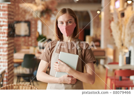 Horizontal waist up portrait shot of young blond woman wearing apron and protective mask working in cafe holding digital tablet looking at camera Horizontal waist up portrait shot of young blond woman wearing apron and protective mask working in cafe holding digital tablet looking at camera 88079466