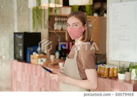 Horizontal medium portrait shot of young woman wearing apron and protective mask starting workday in cafe holding digital tablet looking at camera Horizontal medium portrait shot of young woman wearing apron and protective mask starting workday in cafe holding digital tablet looking at camera 88079468