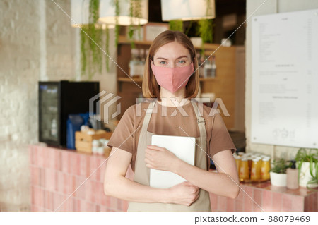 Horizontal medium portrait of young waitperson wearing apron and protective mask holding digital tablet looking at camera Horizontal medium portrait of young waitperson wearing apron and protective mask holding digital tablet looking at camera 88079469