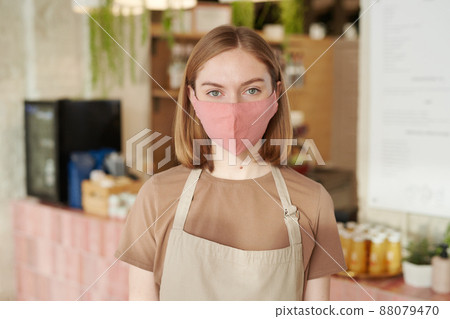 Horizontal medium close-up portrait of young woman wearing apron and protective mask starting workday in cafe looking at camera Horizontal medium close-up portrait of young woman wearing apron and protective mask starting workday in cafe looking at camera 88079470