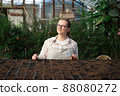 Young woman stands near table with seedlings in large greenhouse and holds rake and shovel in her hands. Gardening, seedlings, sales 88080272