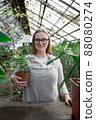 Young woman stands near table with flowers in the greenhouse and holds pot with green flower. Gardening, seedlings, sale 88080274