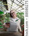 Young woman stands near table with flowers in the greenhouse and holds pot with green flower. Gardening, seedlings, sale 88080275