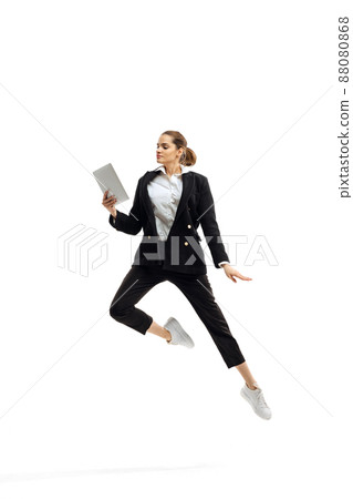 Dynamic portrait of young businesswoman in black suit jumping with tablet, reading isolated over white studio background 88080868