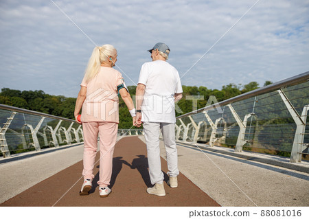 Positive senior woman and man walk holding hands along footbridge after training Positive senior woman and man walk holding hands along footbridge after training 88081016