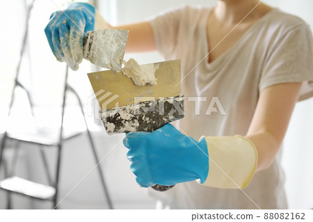 Young female worker preparing putty for the wall repair. Close-up view of spreading plaster on metal spatula. Do it yourself. 88082162