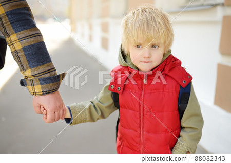 Man with little boy are walking hand in hand along city street. Frustrated child does not want to go to kindergarten or school. Single parent. Father. 88082343