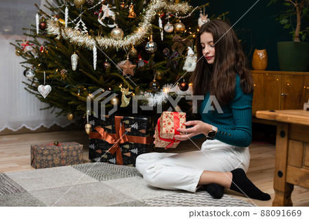 A girl sits by the Christmas tree in the living room with a gift in her hand. 88091669