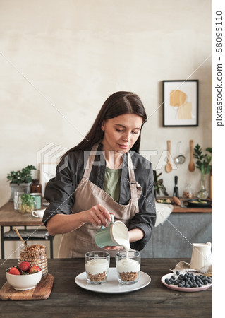 Serious brunette woman in apron leading healthy lifestyle standing at dining table and making granola with yogurt in kitchen 88095110