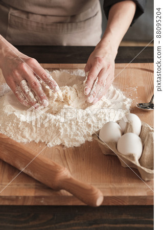Close-up of unrecognizable woman in apron standing at kitchen counter and mixing egg with flour while making dough on wooden board Close-up of unrecognizable woman in apron standing at kitchen counter and mixing egg with flour while making dough on wooden board 88095204