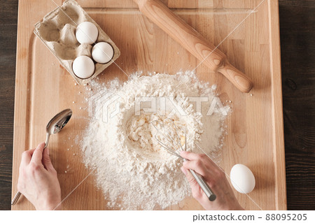 Above view of unrecognizable woman mixing flour and egg with whisk on wooden board with rolling pin 88095205