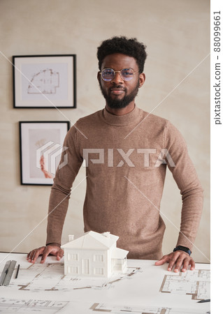 Portrait of content young African-American building architect in glasses standing at table with floor plan and D model of house Portrait of content young African-American building architect in glasses standing at table with floor plan and D model of house 88099661