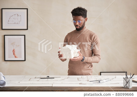 Serious young black architect with beard standing at table with floor plan and analyzing D model of house in office 88099666