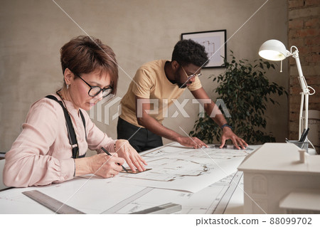 Concentrated mature woman in glasses sitting at table and creating drawing for architectural project together with African-American intern Concentrated mature woman in glasses sitting at table and creating drawing for architectural project together with African-American intern 88099702