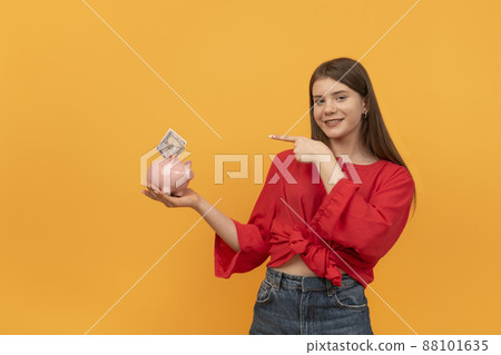 Young woman holds piggy bank and points with finger at it. Concepts of saving money and investments. Young woman holds piggy bank and points with finger at it. Concepts of saving money and investments. 88101635