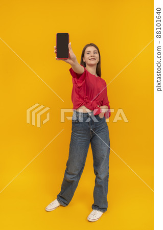 Teenage girl in red blouse holds phone with blank screen. Full-length portrait of girl with smartphone on yellow background. Copy space. Mock up. 88101640
