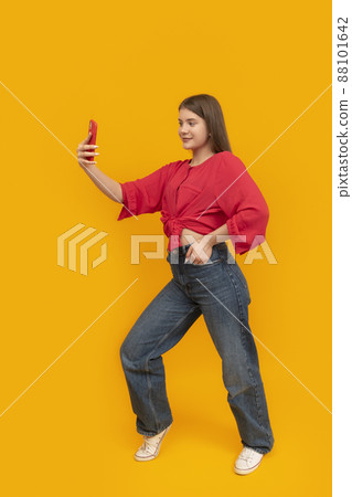 Portrait of teenage girl in red T-shirt and jeans with phone in the studio on yellow background. Young girl takes selfie or shoots stories for social networks. Portrait of teenage girl in red T-shirt and jeans with phone in the studio on yellow background. Young girl takes selfie or shoots stories for social networks. 88101642
