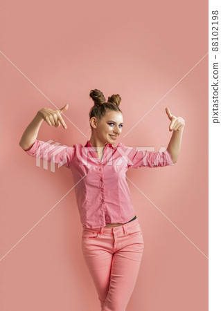 Full-length portrait of young woman in pink shirt and jeans with two bun hairstyles on pink background. Girl points her fingers down. 88102198
