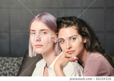 Calm young brown-haired woman leaning on shoulder of girlfriend while embracing her from back in bedroom 88104326