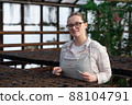 Young woman stands near table with seedlings in large greenhouse and holds rake and shovel in her hands. Gardening, seedlings, sales 88104791