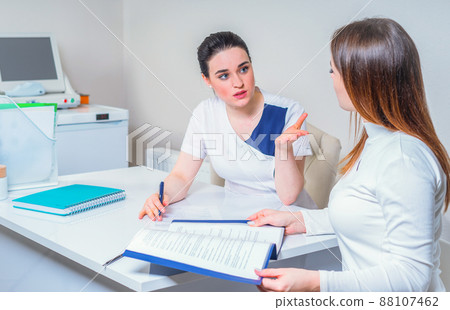 Young female doctor sits at her desk and chats to female patient while showing her test results Young female doctor sits at her desk and chats to female patient while showing her test results 88107462