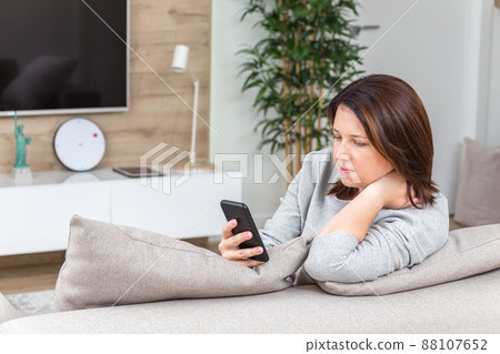 Young woman looking at her smart phone and smiling while sitting on the couch 88107652