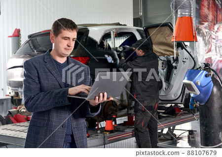 Manager holding laptop while standing near car in auto repair shop with mechanic repairing car on background 88107679