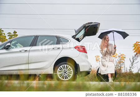 Beautiful woman on road near her white car with punctured car tire. Female driver calling car service for help, holding umbrella, sitting on spare wheel. Beautiful woman on road near her white car with punctured car tire. Female driver calling car service for help, holding umbrella, sitting on spare wheel. 88108336