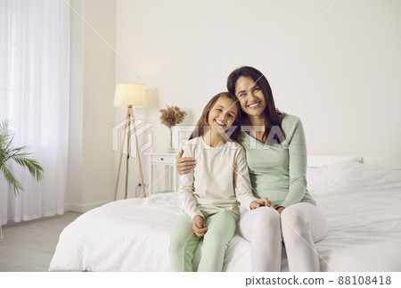 Family portrait of smiling mother and child at home. Happy beautiful mum and daughter in comfortable white and pastel green lounge homewear sitting together on cosy, warm, big, wide bed in bedroom Family portrait of smiling mother and child at home. Happy beautiful mum and daughter in comfortable white and pastel green lounge homewear sitting together on cosy, warm, big, wide bed in bedroom 88108418