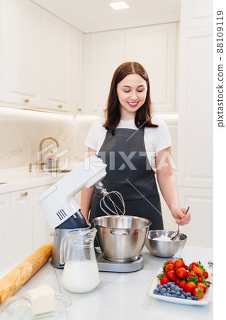 A young woman cook adds a spoonful of flour to prepare dough in a mixer bowl. A young woman cook adds a spoonful of flour to prepare dough in a mixer bowl. 88109119