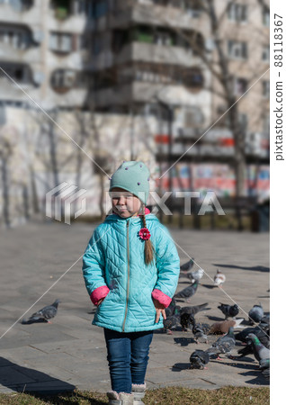 A girl in a blue jacket poses outside in front of a flock of city pigeons. A five-year-old child with long pigtails. Blurred image of bird food in the background. Daytime. Springtime. A girl in a blue jacket poses outside in front of a flock of city pigeons. A five-year-old child with long pigtails. Blurred image of bird food in the background. Daytime. Springtime. 88118367