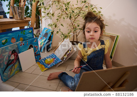 Adorable preschooler girl enjoying learning painting, sitting on the floor at a wooden easel against the background of colorful joyful children painted images. Art class, creativity and education 88132186