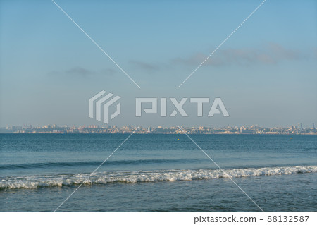 Sea waves running on a sandy shore on a clear sunny day. City in the background. Sea waves running on a sandy shore on a clear sunny day. City in the background. 88132587