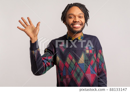 Hi you. Positive friendly african man with dreadlocks and beard waving hand greeting you with toothy smile on face, nice to meet, dating. Indoor studio shot isolated on gray background 88133147