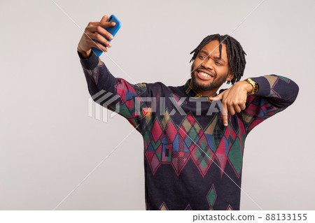Handsome self confident african male blogger with dreadlocks showing subscribe gesture looking at camera of smartphone with toothy smile. Indoor studio shot isolated on gray background 88133155