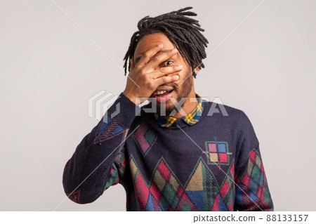 Curious nosy african guy with dreadlocks spying through hole in fingers closing eyes with arm and smiling, peeking. Indoor studio shot isolated on gray background 88133157