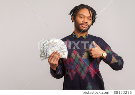 Pleased self confident african man with beard and dreadlocks showing thumbs up gesture holding fan of dollars, cashback, crediting. Indoor studio shot isolated on gray background Pleased self confident african man with beard and dreadlocks showing thumbs up gesture holding fan of dollars, cashback, crediting. Indoor studio shot isolated on gray background 88133256