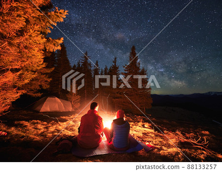 Back view of male and female best friends resting near night bonfire during mountaineering vacations for visiting picturesque nature environment, husband and wife have wild honeymoon for trekking Back view of male and female best friends resting near night bonfire during mountaineering vacations for visiting picturesque nature environment, husband and wife have wild honeymoon for trekking 88133257