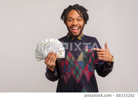 Extremely satisfied cheerful african man with dreadlocks pointing finger on dollars fan in his hand, money crediting, easy loan. Indoor studio shot isolated on gray background Extremely satisfied cheerful african man with dreadlocks pointing finger on dollars fan in his hand, money crediting, easy loan. Indoor studio shot isolated on gray background 88133282