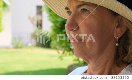 portrait of pensive mature woman aged 55-60 in sun-protective straw hat against green garden 88133350