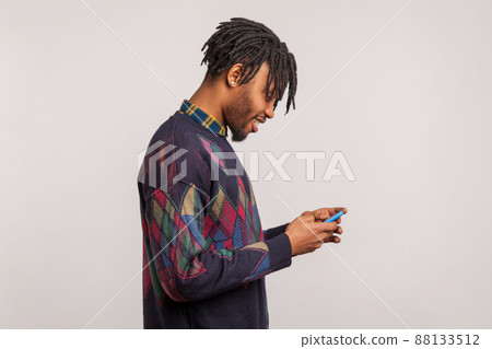 Profile portrait of happy trendy african guy with dreadlocks in sweatshirt using smartphone and smiling, pleased satisfied with connection quality. Indoor studio shot isolated on gray background 88133512