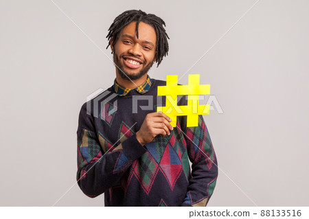 African man with dreadlocks and toothy smile on his face holding hashtag symbol, recommending popular topics, internet trends. Indoor studio shot isolated on gray background African man with dreadlocks and toothy smile on his face holding hashtag symbol, recommending popular topics, internet trends. Indoor studio shot isolated on gray background 88133516