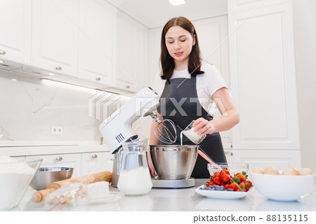 A women cook adds a milk to prepare dough or cream in a mixer bowl. A women cook adds a milk to prepare dough or cream in a mixer bowl. 88135115
