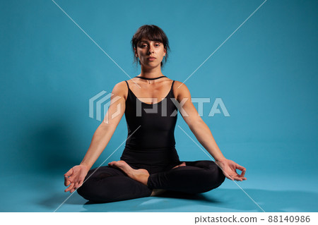 Concentrated trainer meditating during pilates workout sitting in lotus position on yoga mat practicing spiritual meditation. Gymnast woman training body muscles in studio with blue background Concentrated trainer meditating during pilates workout sitting in lotus position on yoga mat practicing spiritual meditation. Gymnast woman training body muscles in studio with blue background 88140986